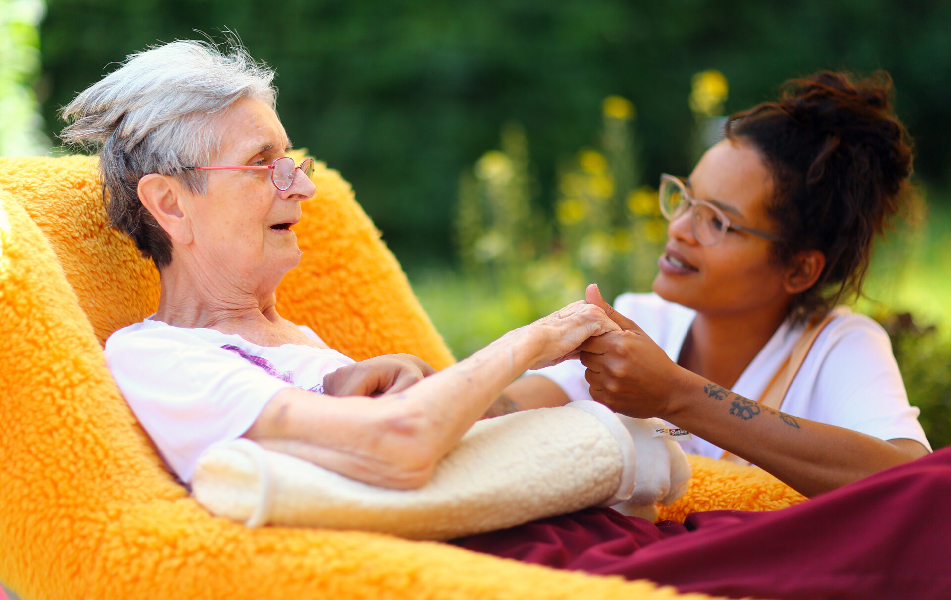 Eine schwer an Demenz erkrankte Frau sitzt in einem gelben Sessel. Eine junge Pflegerin hält ihre Hand.