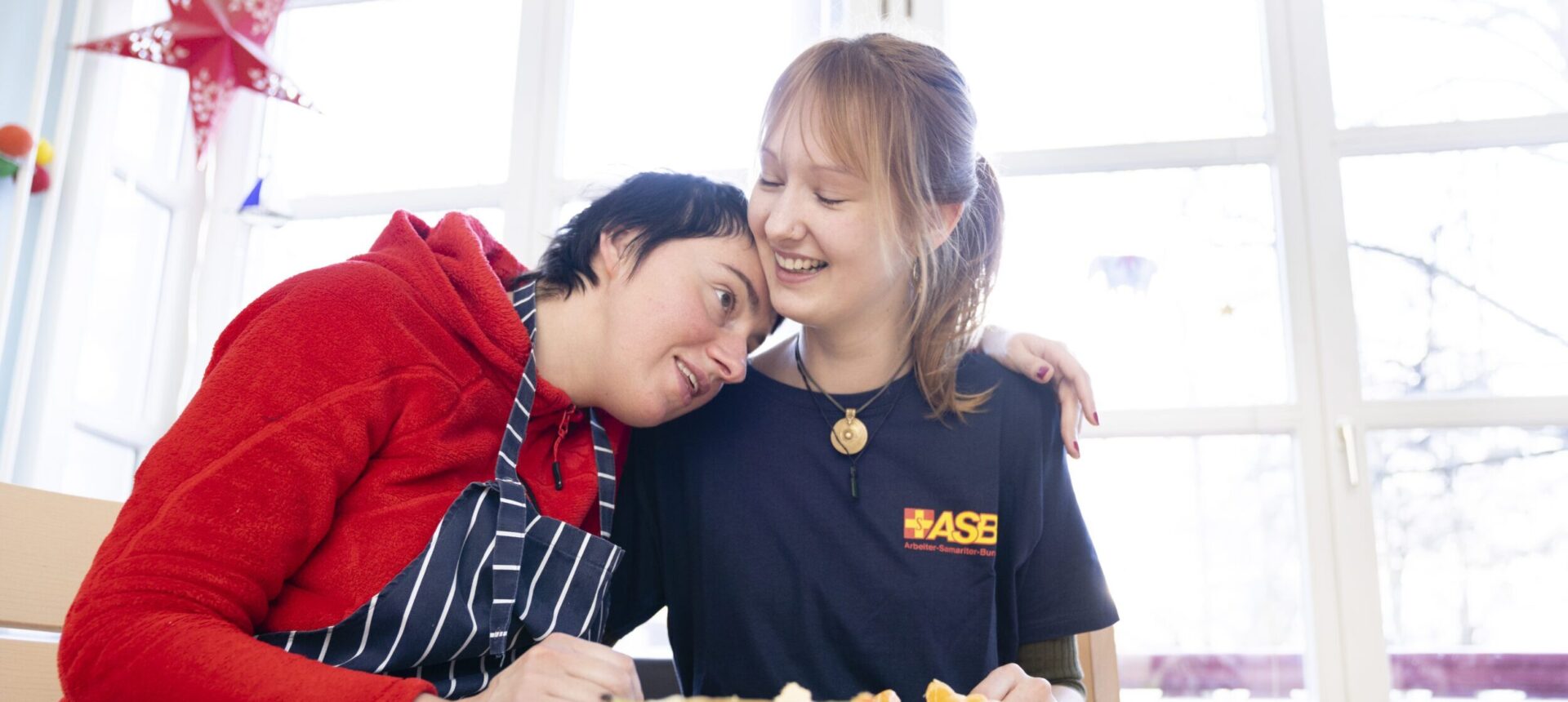Eine Frau mit geistiger Behinderung umarmt in der Küche eine ASB-Mitarbeiterin. Beide waren gerade dabei, gemeinsam Obstsalat vorzubereiten.