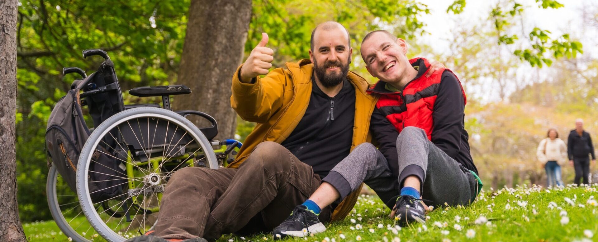 Ein Heilerziehungspfleger und ein junger Mann sitzen im Park auf einer Wiese. Im Hintegrund steht ein Rollstuhl.