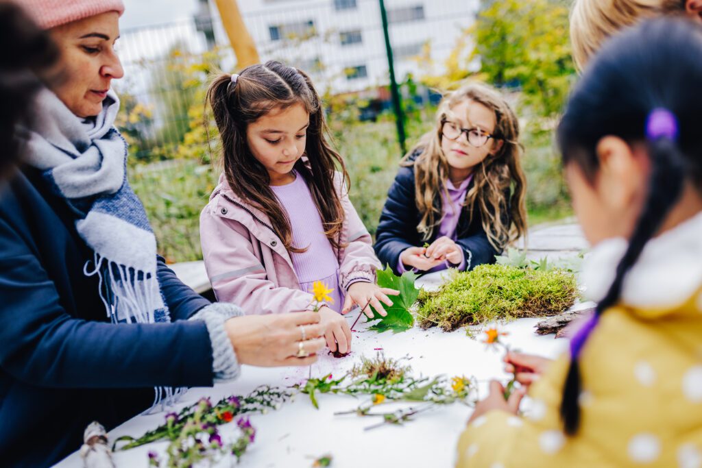 Einige Kinder stehen an einem Tisch und schauen sich mit einer Frau verschiedene Blumen, Blätter und Moos an.