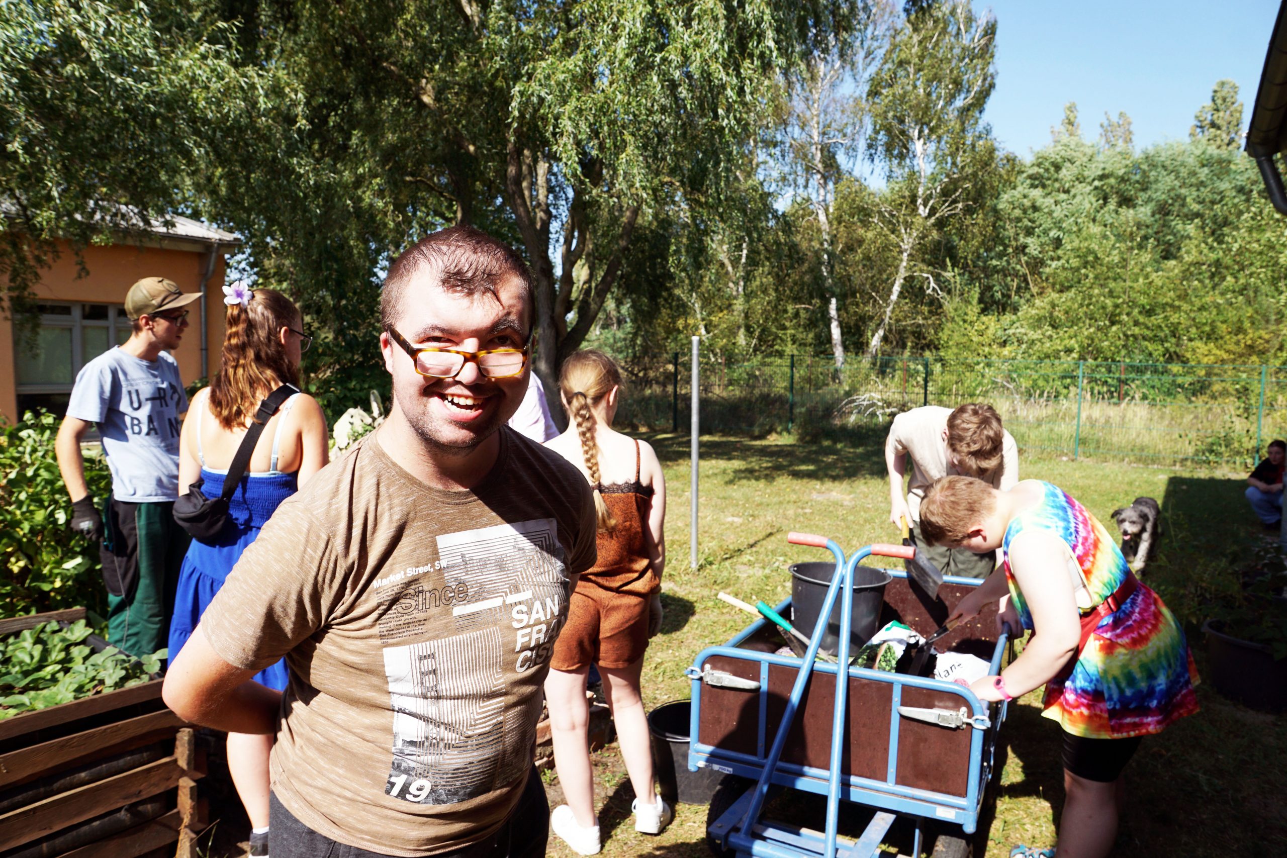 Ein junger Herr lacht in die Kamera. Hinter ihnen stehen einige junge Menschen, die Gartenarbeit verrichten.