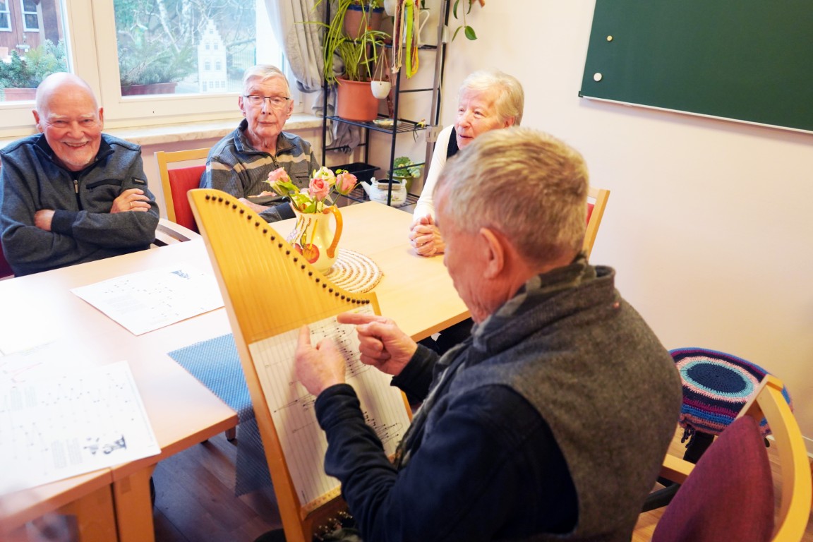 Einige Senioren sitzen ringsrum an einem Tisch. Einer von ihnen spielt die Veeh-Harfe, ein Zupfinstrument. Die anderen schauen zu, einer lacht in die Kamera.