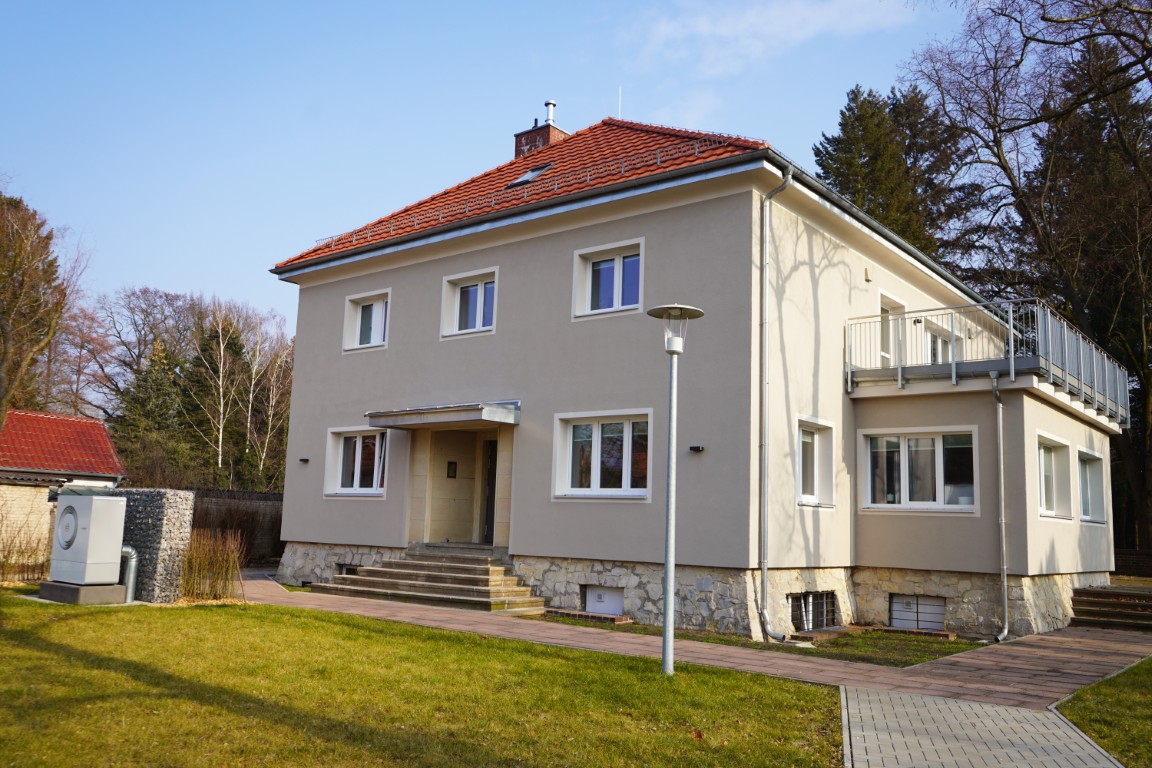 Das Haus Lichtblick von außen. Eine beige Fassade mit einem rot-orangenen Dach und einem Anbau an der rechten Seite mit Terasse. Davor liegt ein Weg und eine grüne Wiese.