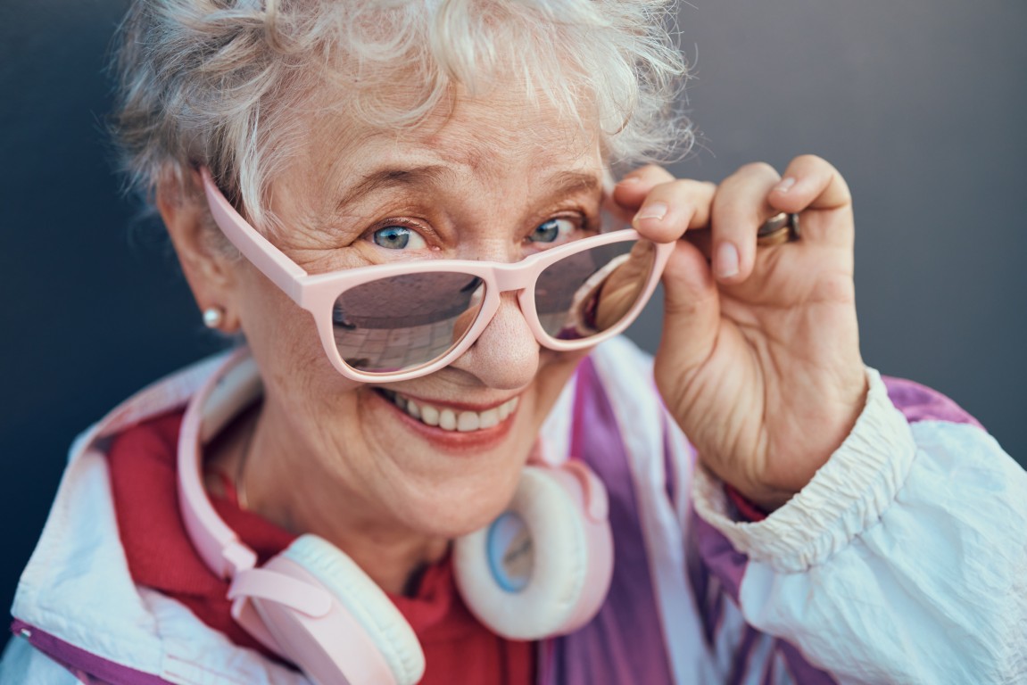 Eine ältere Dame die grinsend über ihre Sonnenbrille in die Kamera schaut. Sie trägt bunte, sportliche Kleidung und Kopfhörer um den Nacken.