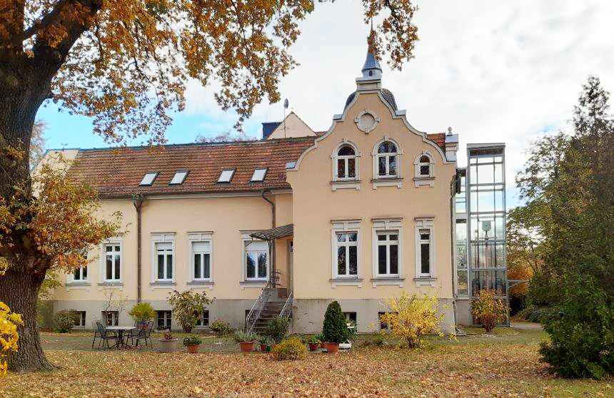 Ein Haus mit einer geschwungenen Front mit Ornamenten. An der rechten Seite des Hauses befindet sich ein gläserner Fahrstuhl.