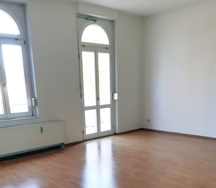 Ein leeres Zimmer. Auf der rechten Seite ist ein Fenster mit einem Bogen als Rahmen, darunter eine Heizung. Daneben eine Glastür, ebenfalls mit einem Bogen als Rahmen.