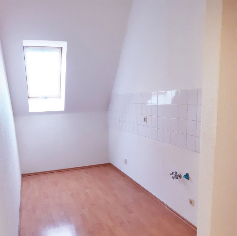 Ein leeres Zimmer im Dachgeschoss mit einer Fliesenwand auf der rechten Seite und einem Fenster, durch das Licht fällt.