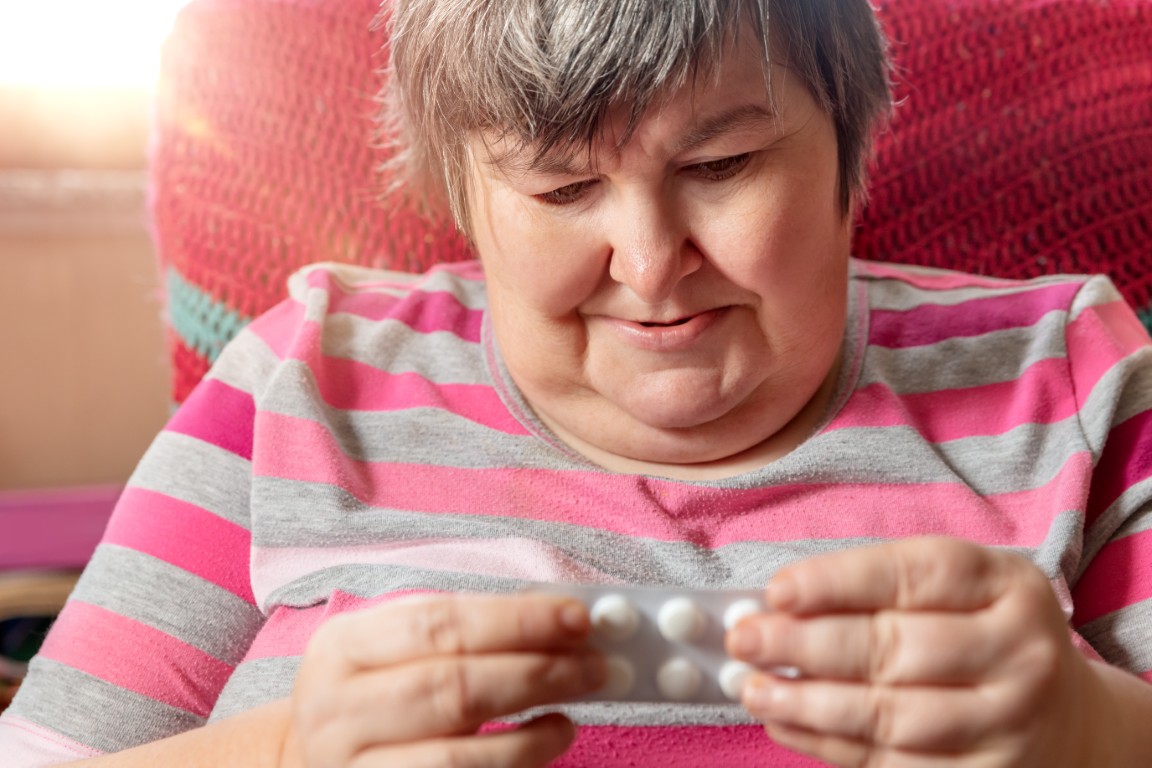 Eine Dame sitzt auf einem Sessel und schaut nachdenklich auf eine Tabletten-Packung, die sie in der Hand hält.