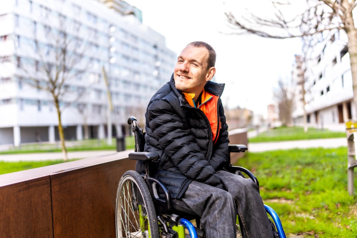 Ein junger Mann sitzt in einem Rollstuhl in einem Park und lächelt.